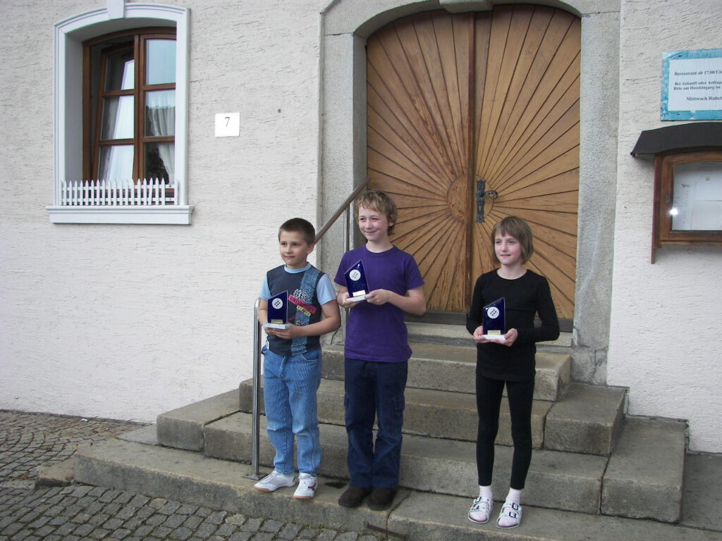 Tři děti držící trofeje stojí na kamenných schodech před budovou s dřevěnými dveřmi a oknem s bílým plotem.