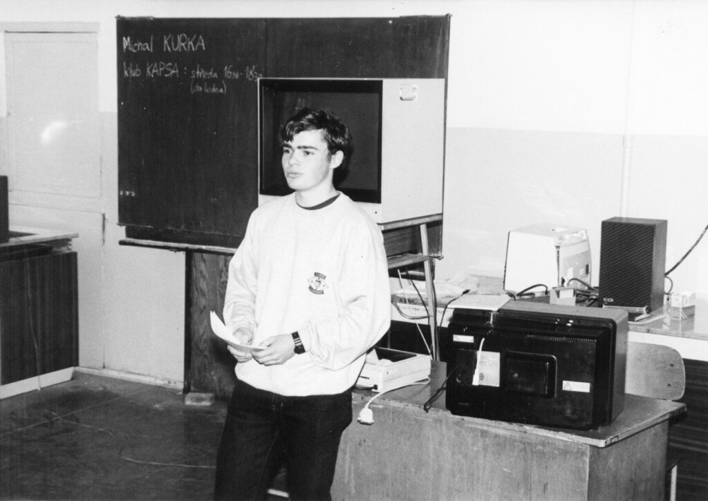 Černobílá fotografie osoby stojící ve třídě u tabule zobrazující "Michal KURKA" a "klub KAPSA : streda 16:30-18:30 (do ledna)," držící papíry, s historickou televizí na stole.