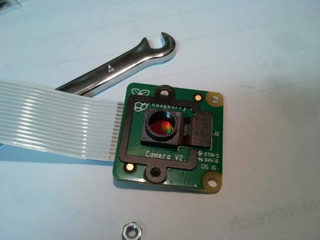 Raspberry Pi Camera V2 modul s plochým kabelem, kovovým klíčem a matičkou na bílém povrchu.