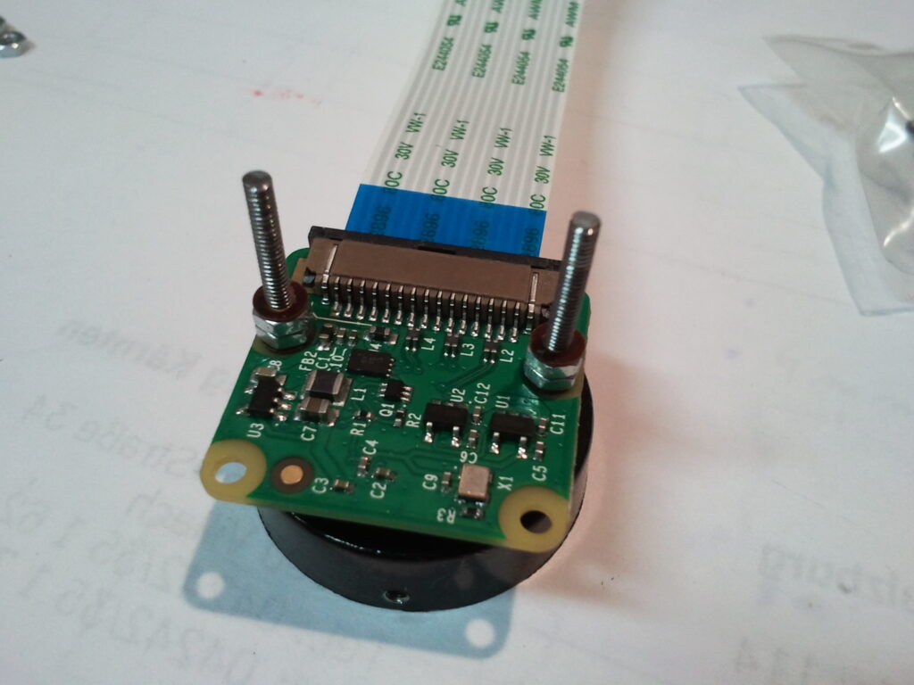 Zelená deska s tištěnými obvody kamery s elektronickými součástmi, dvěma montážními šrouby a černým válcovým objektivem pod vícežilovým kabelem.