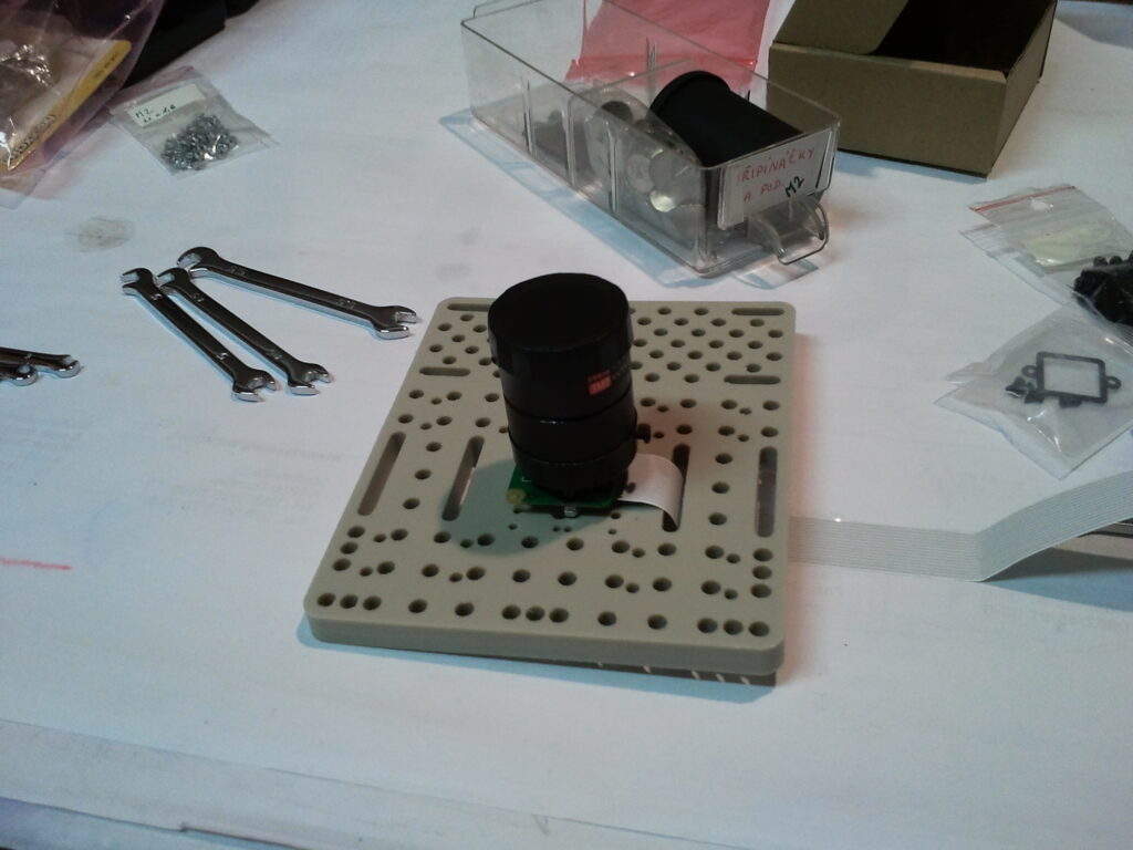 Deska s objektivem kamery, kovovými klíči a roztříděným spojovacím materiálem na pracovním stole.