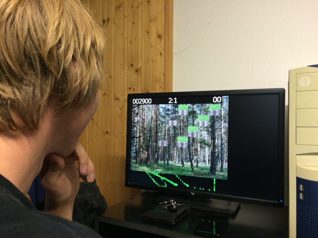 Student s blond vlasy kouká na monitor zobrazující lesní scénu se zelenými bloky a číselné údaje, v pozadí dřevem obložený kout předsíňky s botníky.