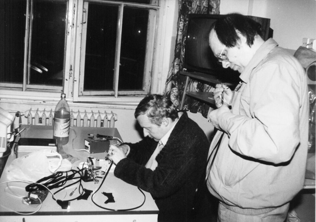 Dva muži pracují na elektronickém zařízení u stolu v místnosti s oknem, radiátorem a televizí.