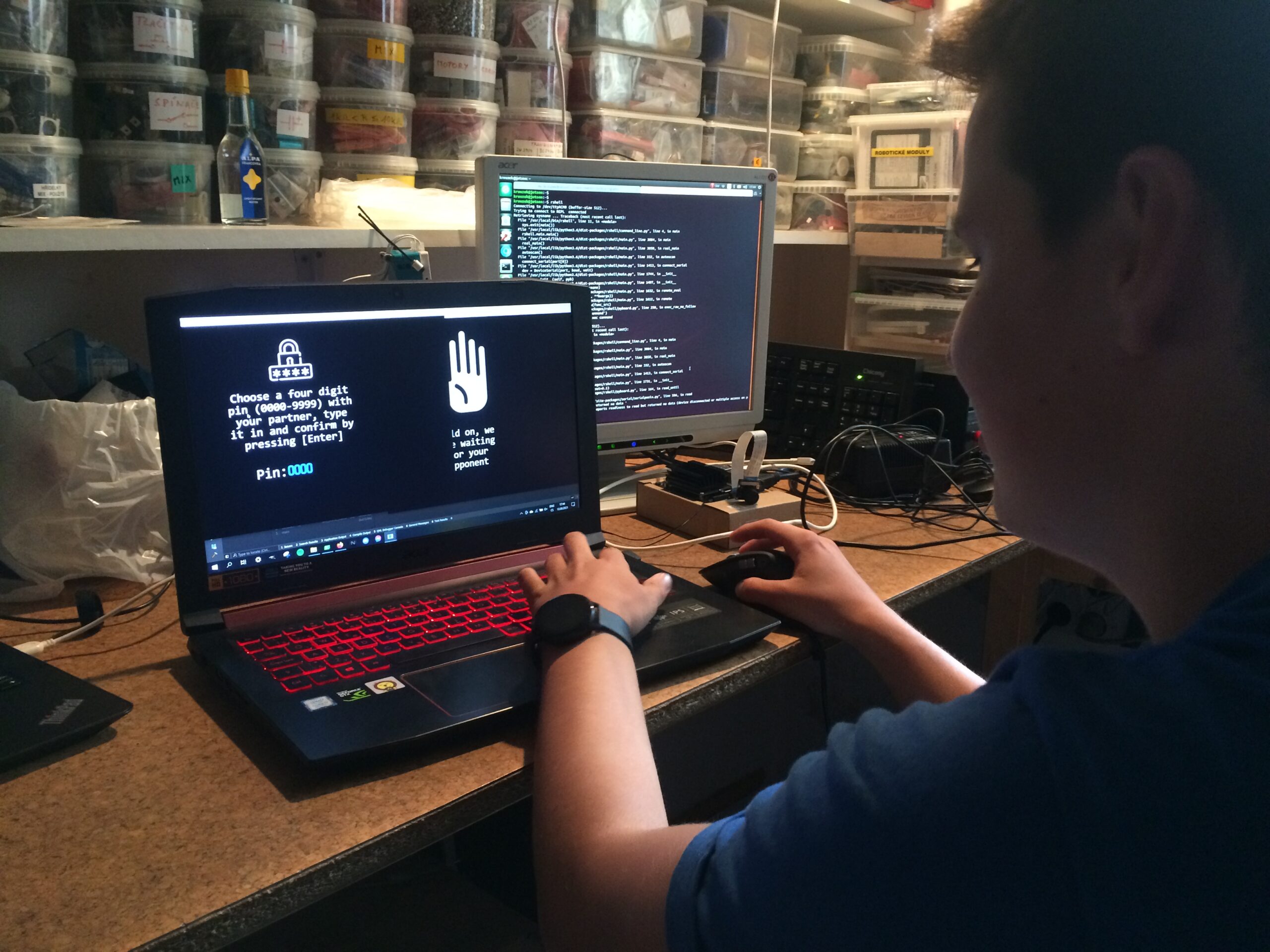 Student, který používá notebook s červeně osvětlenou klávesnicí, displej zobrazuje výzvu k zadání PIN a vedle monitor na stole zobrazující kód, v pracovním prostoru se stohovanými skladovacími kontejnery.