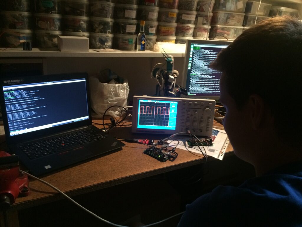Student pracuje u stolu s notebookem s nápisem "KAPSA Robotics" a kódem, monitorem zobrazujícím text, osciloskopem zobrazující hranatý signál a v pozadí je množství krabiček se součástkami na policích.