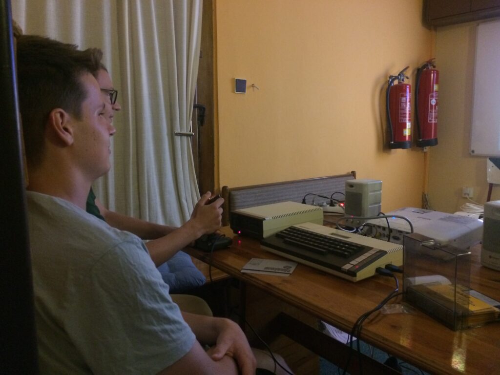 Dva studenti sedí u dřevěného stolu s historickým počítačem Atari, jeden drží joystick. Na stole dále leží disketová mechanika, karabička disket, reproduktory a obraz se proimítá na plátno v pozadí.