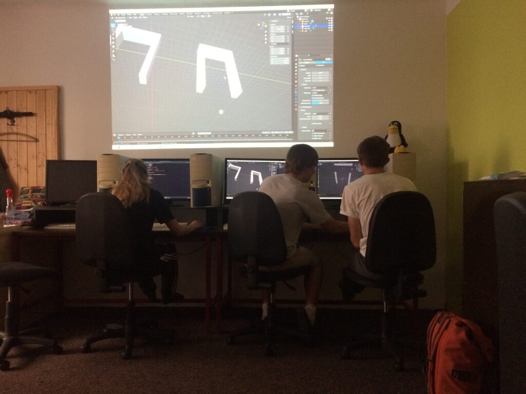 Student pomáhá druhému s modelováním v Blenderu, nad nimi je promítnutý obraz 3D modulu rámu teleportu.