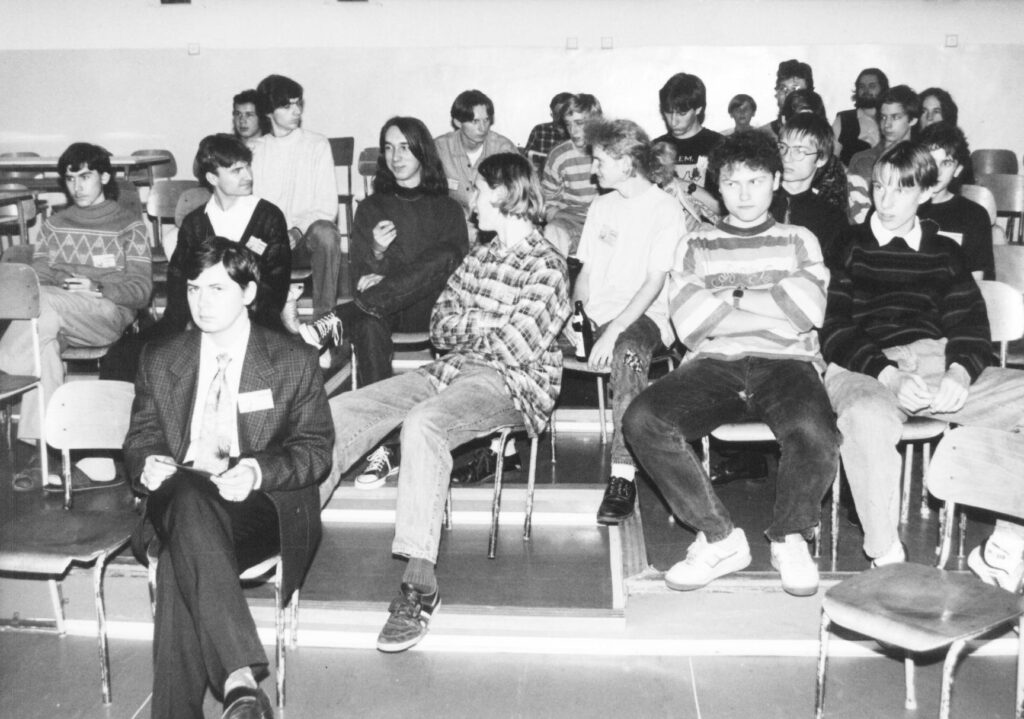 Černobílá fotografie skupiny lidí sedících ve třídě.