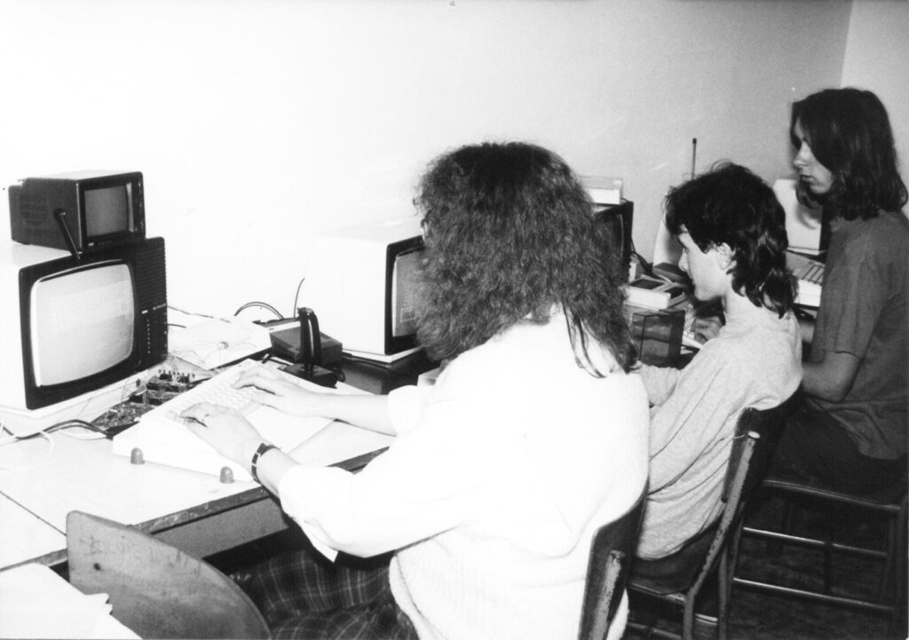 Tři lidé pracují na historických počítačích s CRT monitory v černobílé fotografii.
