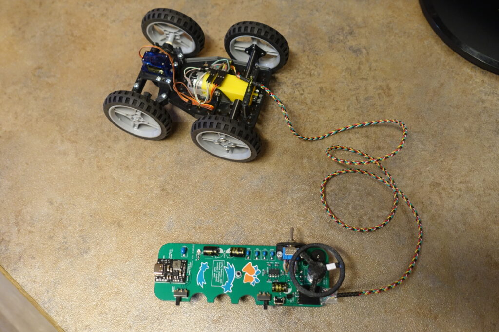 Čtyřkolové robotické auto s černými pneumatikami a bílými ráfky je připojeno vícebarevnými dráty k zelené desce s volantem, elektronickými součástmi a nálepkami ve tvaru ryb.