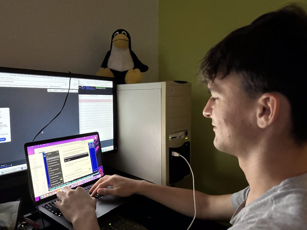 Student píše na notebooku u monitoru, na počítačové skříni sedí plyšový tučňák Tux.