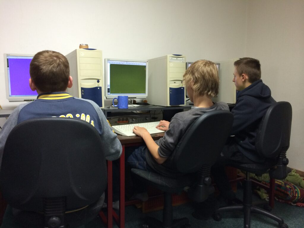Tři děti sedí u počítačů s monitory a modrým hrníčkem v místnosti.