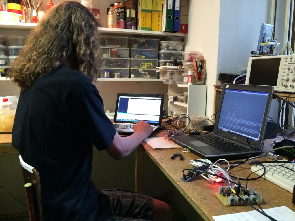 Student s dlouhými kudrnatými vlasy používá laptop u přeplněného stolu s dalším notebookem, elektronickými součástkami a policemi v pozadí.