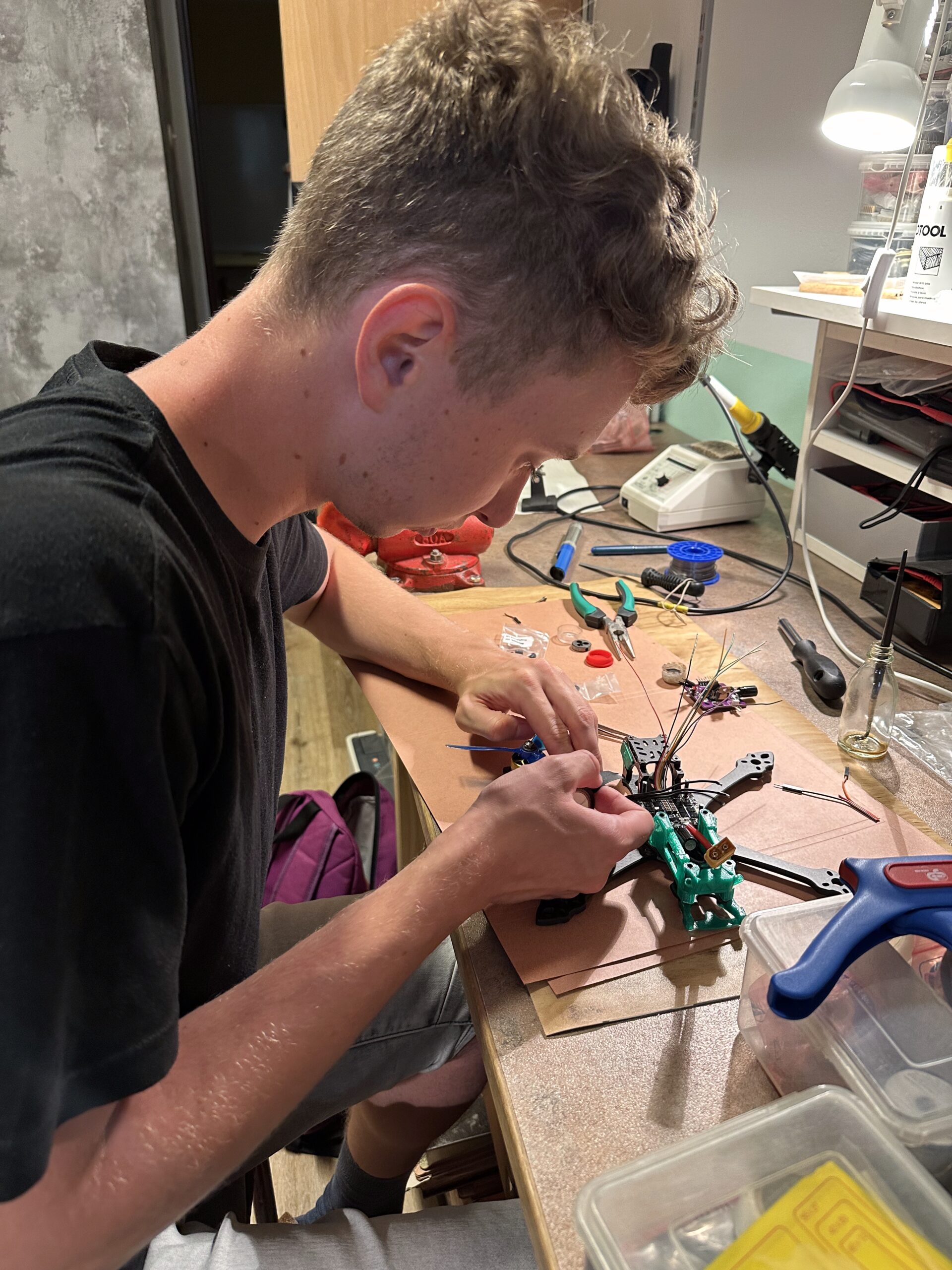 Student v černém tričku montuje elektroniku do rámu dronu na pracovním stole s nástroji a vybavením.