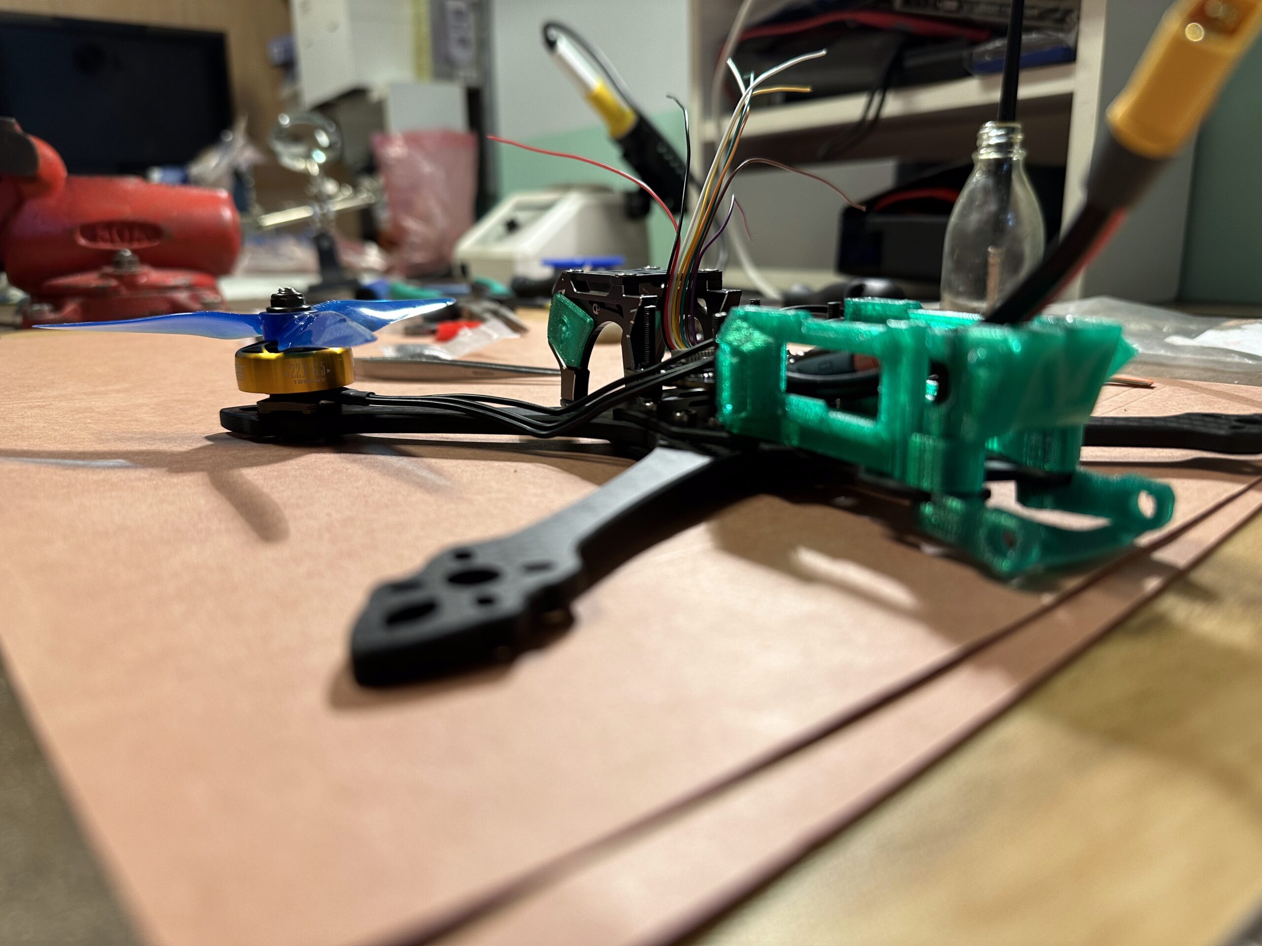 Sestavovaný dron s modrými vrtulemi, zeleným 3D tisknutým rámem a dráty na pracovním stole.