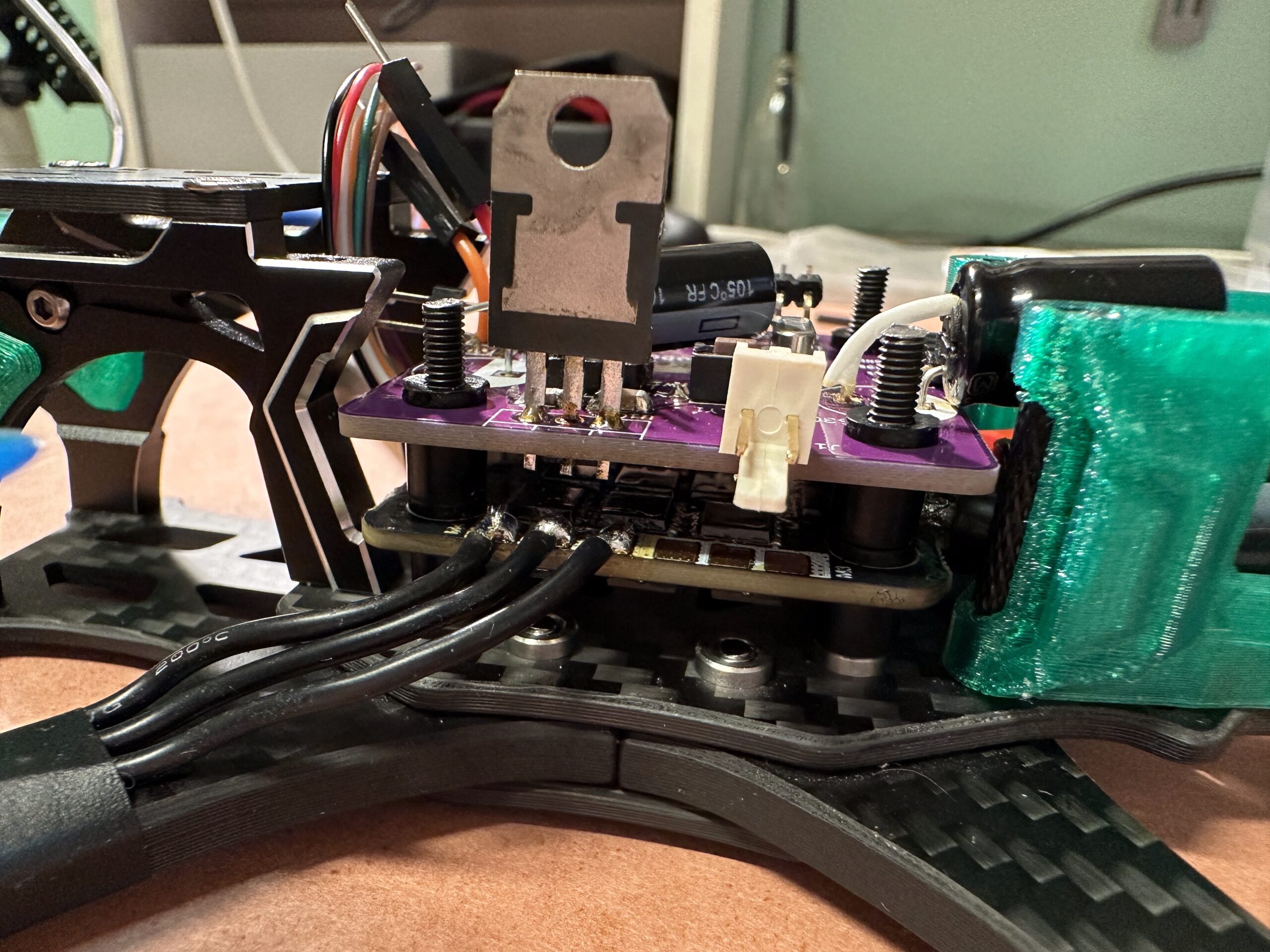 Detail elektronické sestavy s fialovou deskou, dráty, konektory, zeleným plastovým dílem a kovovými částmi na černém rámu dronu.