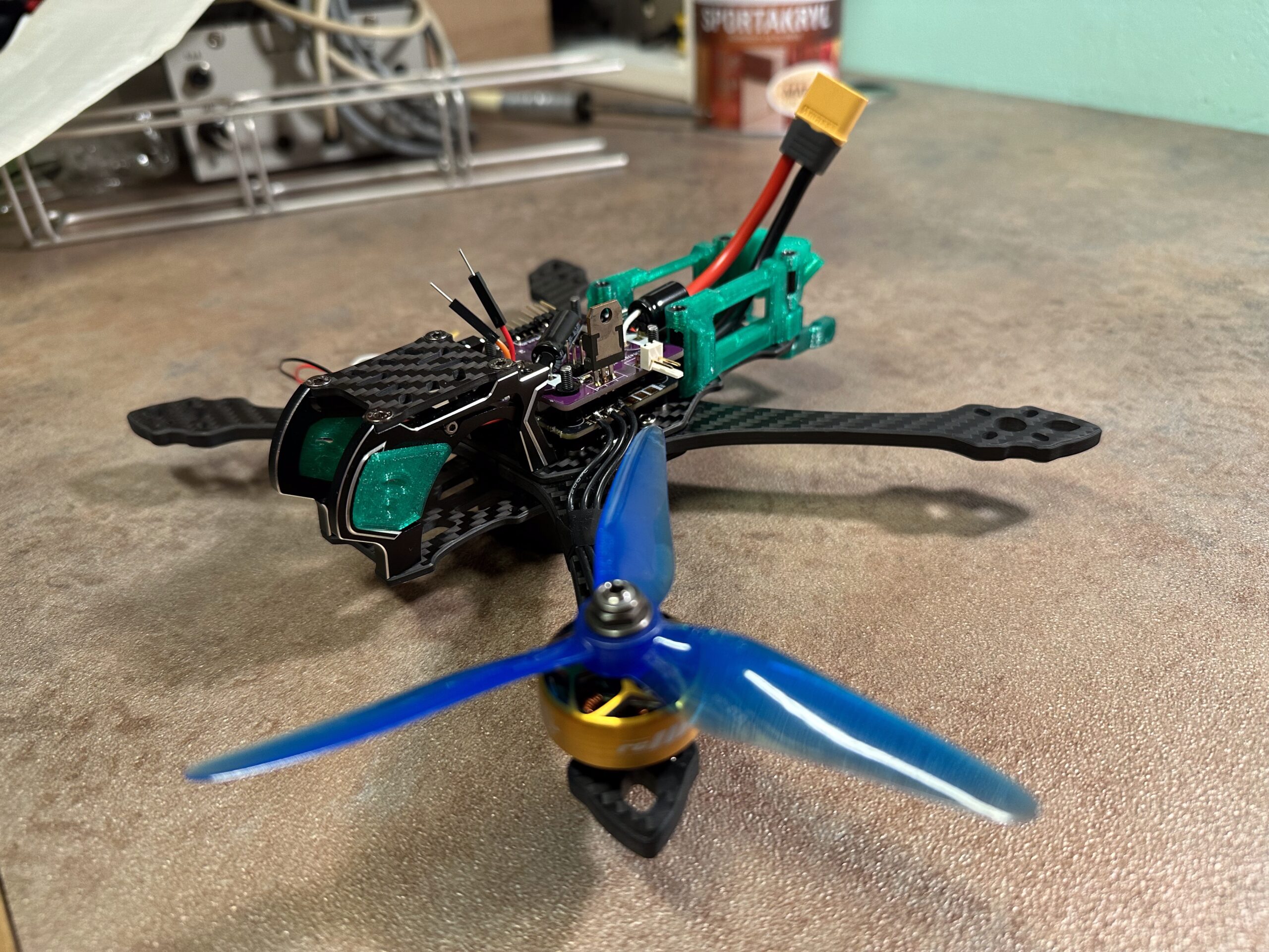 Malý dron s modrými vrtulemi, černými karbonvými rameny a viditelnými elektronickými součástkami.