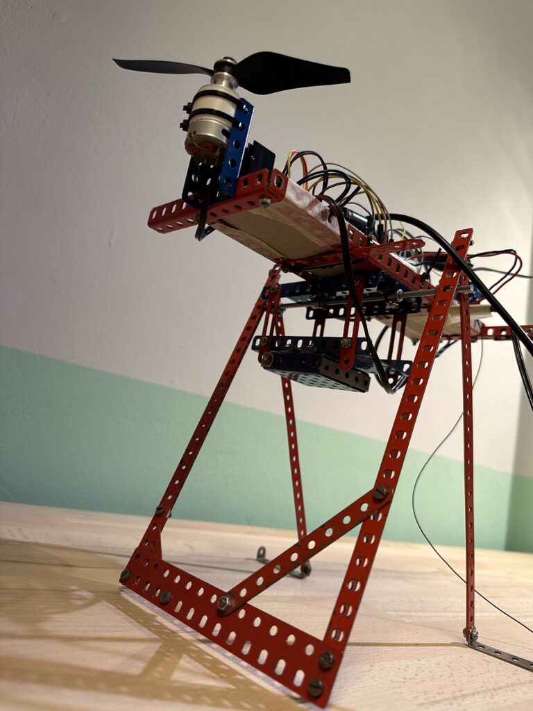 Červená kovová houpačka s vrtulemi na krajích a elektroinstalací na dřevěném stole proti dvoutónové stěně.