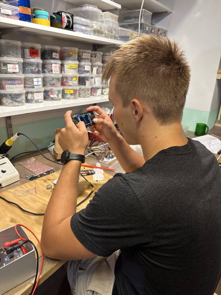 Student drží vyrobenou elektronickou hru s displejem v dílně s policemi označených součástek.