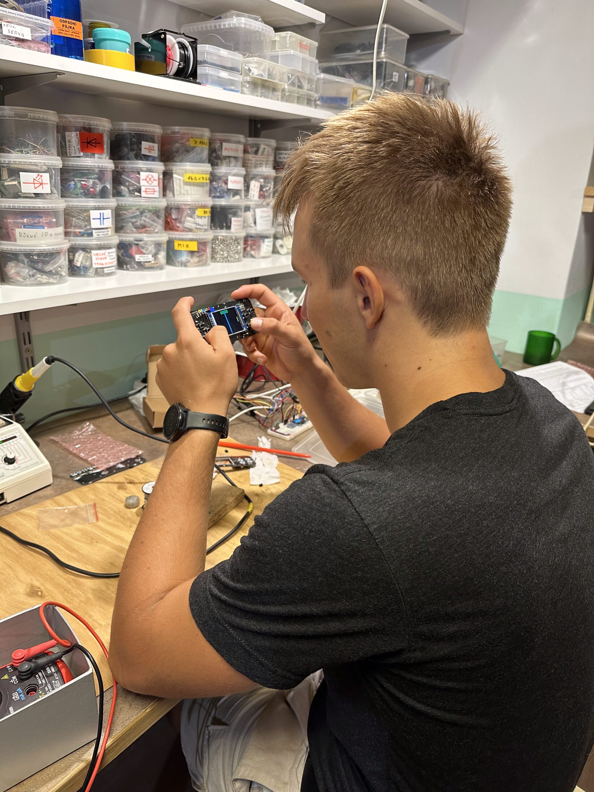 Student drží vyrobenou elektronickou hru s displejem v dílně s policemi označených součástek.