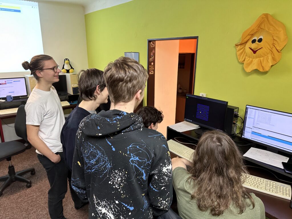 Skupina dětí a studentů u počítačů v místnosti se zelenými stěnami, plyšovým sluníčkem a projektorem.