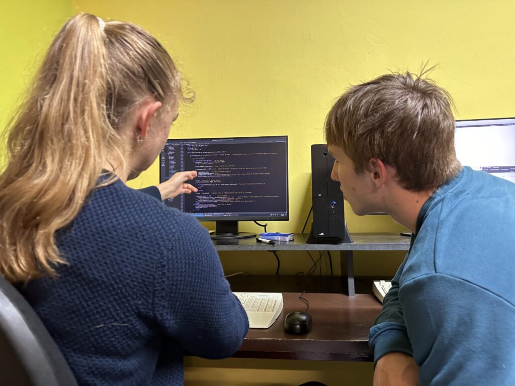 Kluk a holka sledují kód na monitoru u stolu, žlutá stěna v pozadí.