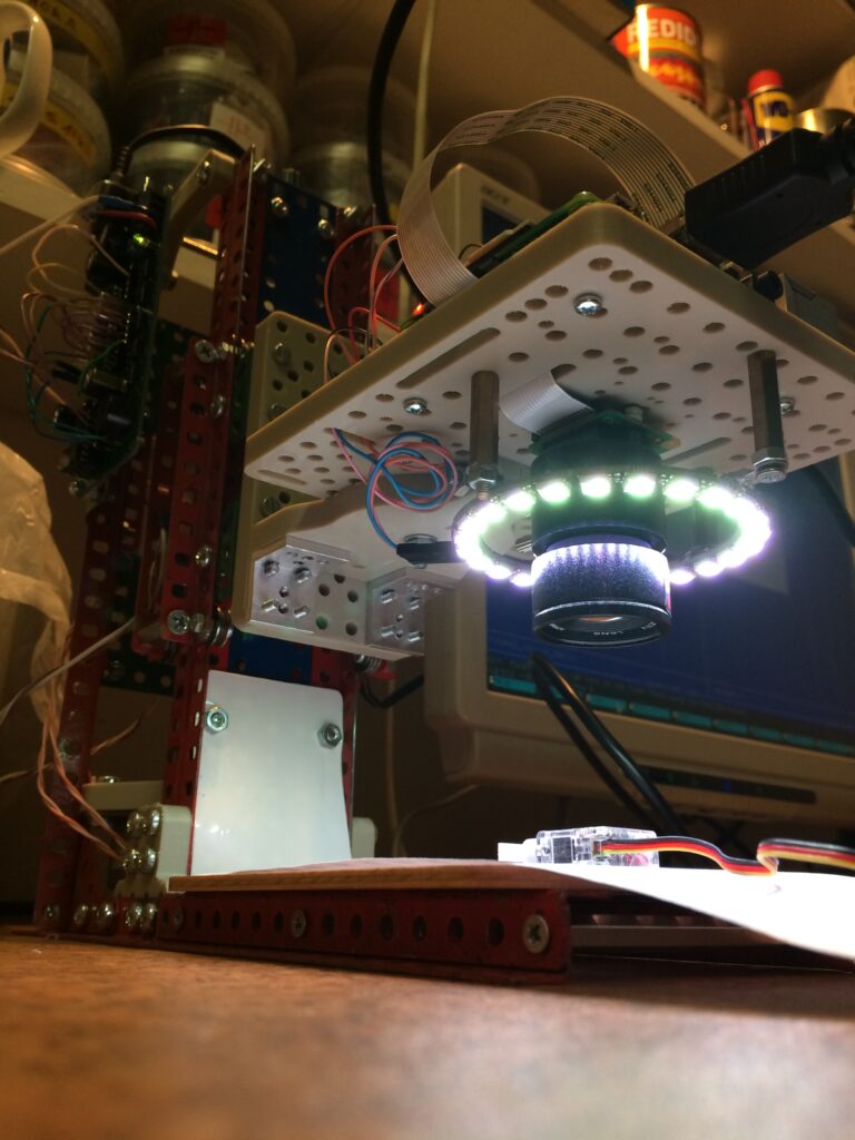Kamera s kruhovým polem osvětlených LED diod namontovaná na perforované desce, obklopené dráty, kovovými lištami a elektronickými součástmi.
