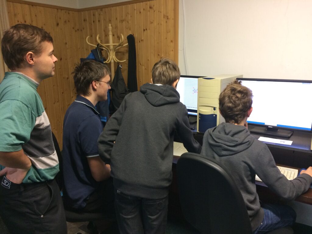 Tři žáci a učitel se shromažďují kolem počítačových monitorů v místnosti s dřevěnými stěnami a věšák na kabáty.