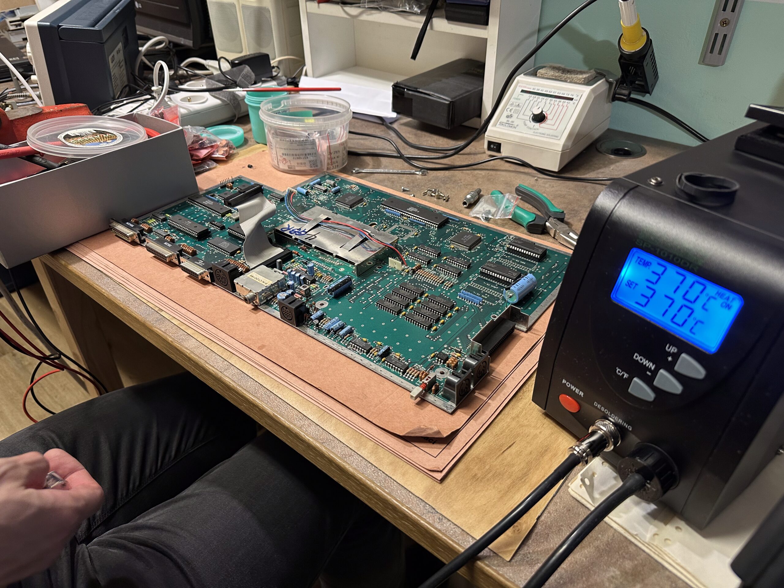 Pracovní stůl se zelenou deskou plošných spojů, pájecí stanicí s 370 °C a různými nástroji pro elektroniku.