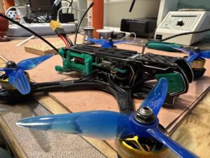 Dron s modrými vrtulemi a zeleným rámem na pracovním stole obklopeném nářadím a páječkou.