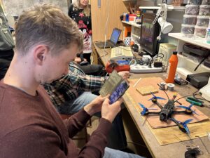 Student drží smartphone zobrazující obraz foceného dronu v elektotechnické dílně s nástroji, elektronikou a poličkami zásob.