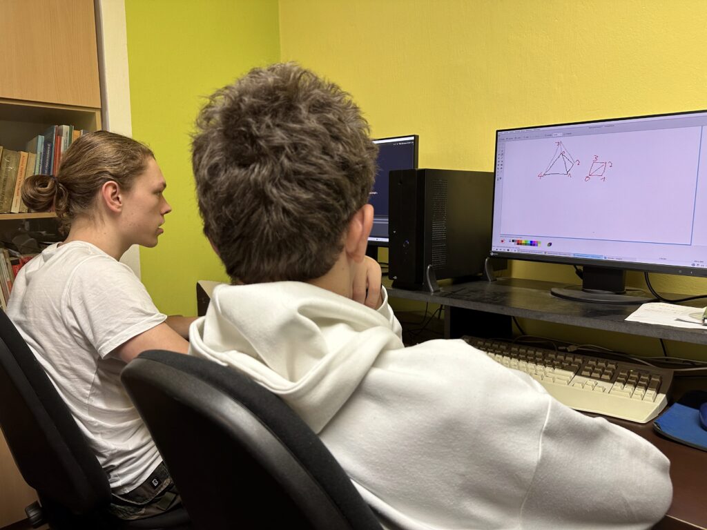 Dva studenti u monitorů zobrazujících náčrtek geometrie pyramidy, v pozadí žlutá stěna a knihovna.