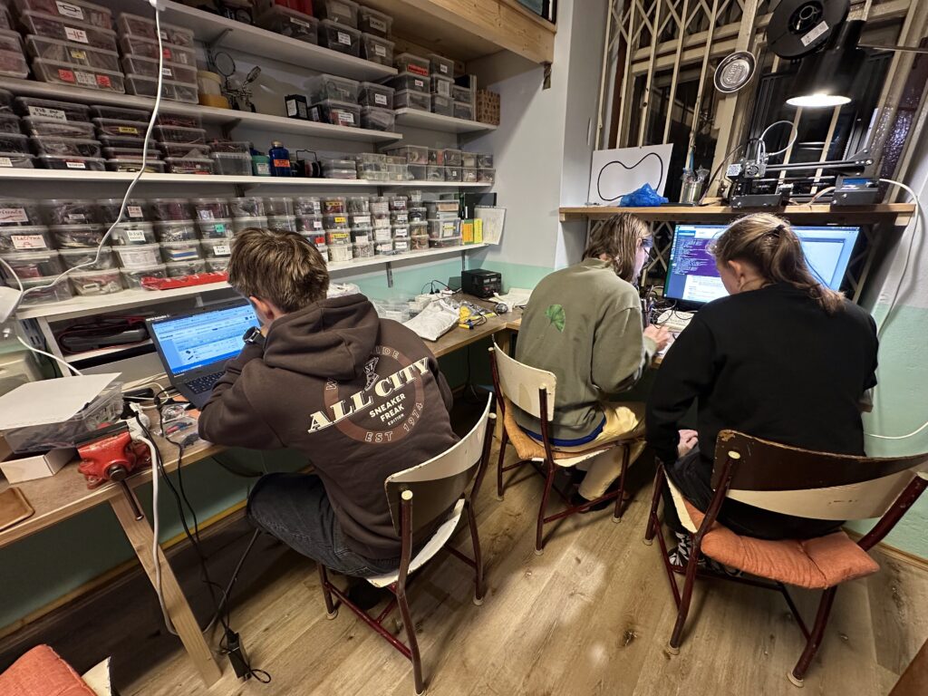 Tři studenti pracují u stolů v dílně s organizovanými zásobníky, nářadím a 3D tiskárnou.