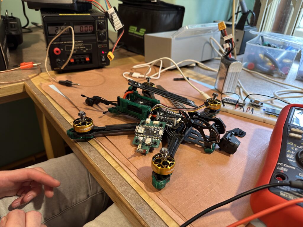 Pracovní stůl s částečně sestaveným dronem, součástkami vč. kamery, multimetrem a pájecí stanicí.