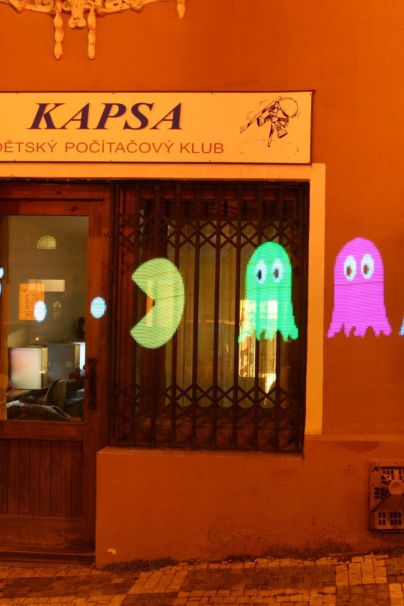 Fasáda s nápisem "KAPSA DĚTSKÝ POČÍTAČOVÝ KLUB" v noci s promítanou zelenou a růžovou grafikou duchů před výlohou s kovovými mřížemi a dřevěnými dveřmi.