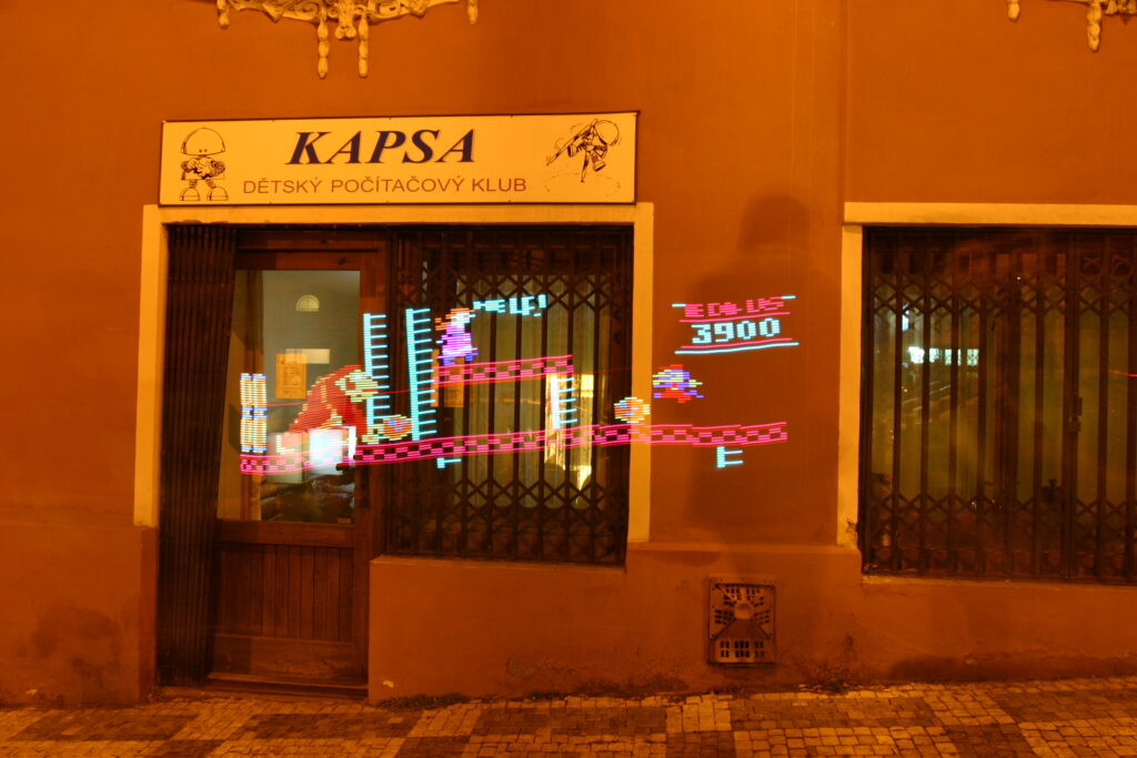 Noční exteriér budovy s nápisem "KAPSA DĚTSKÝ POČÍTAČOVÝ KLUB" a neonovou grafikou pixelové videohry před výlohou.