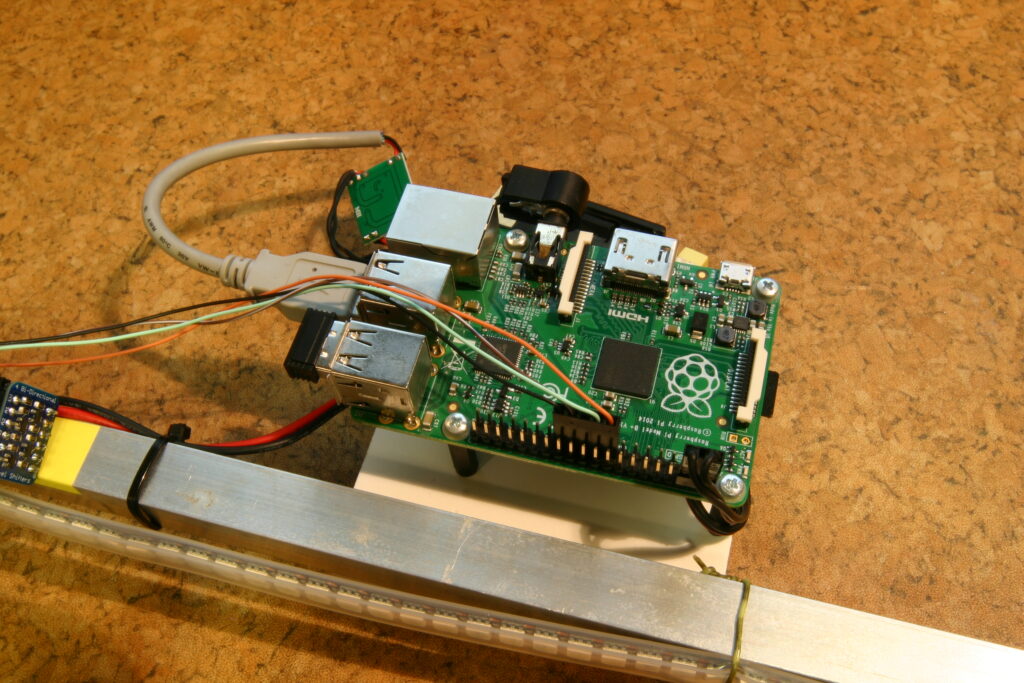 Raspberry Pi deska s připojenými USB kabely, dráty, kovová lišta s LED pásy, a elektronické komponenty na korkovém povrchu.