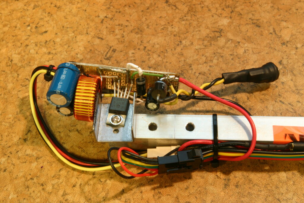 Elektronická deska s modrým kondenzátorem, měděnou cívkou, tranzistorem, rezistory, vícebarevnými dráty, kovovou konzolou a konektorem na korkovém povrchu.