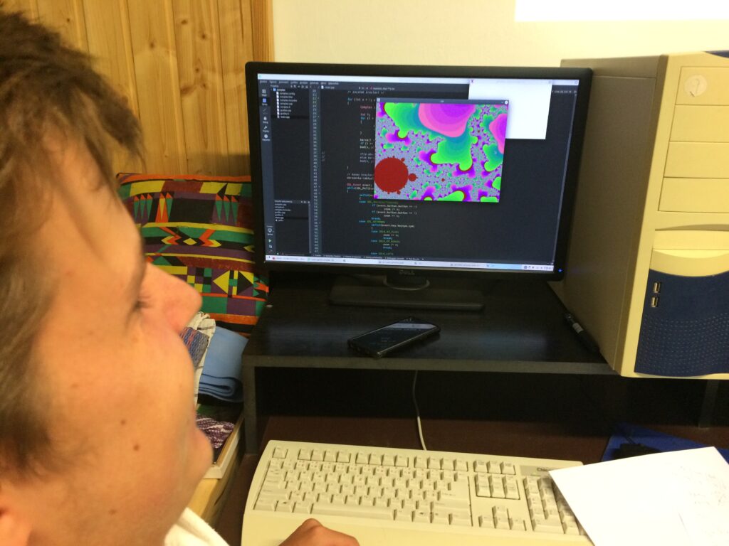 Student u počítače, na obrazovce je zvětšený výřez z Maldenbrotovy množiny a počítasčový kód v pozadí.