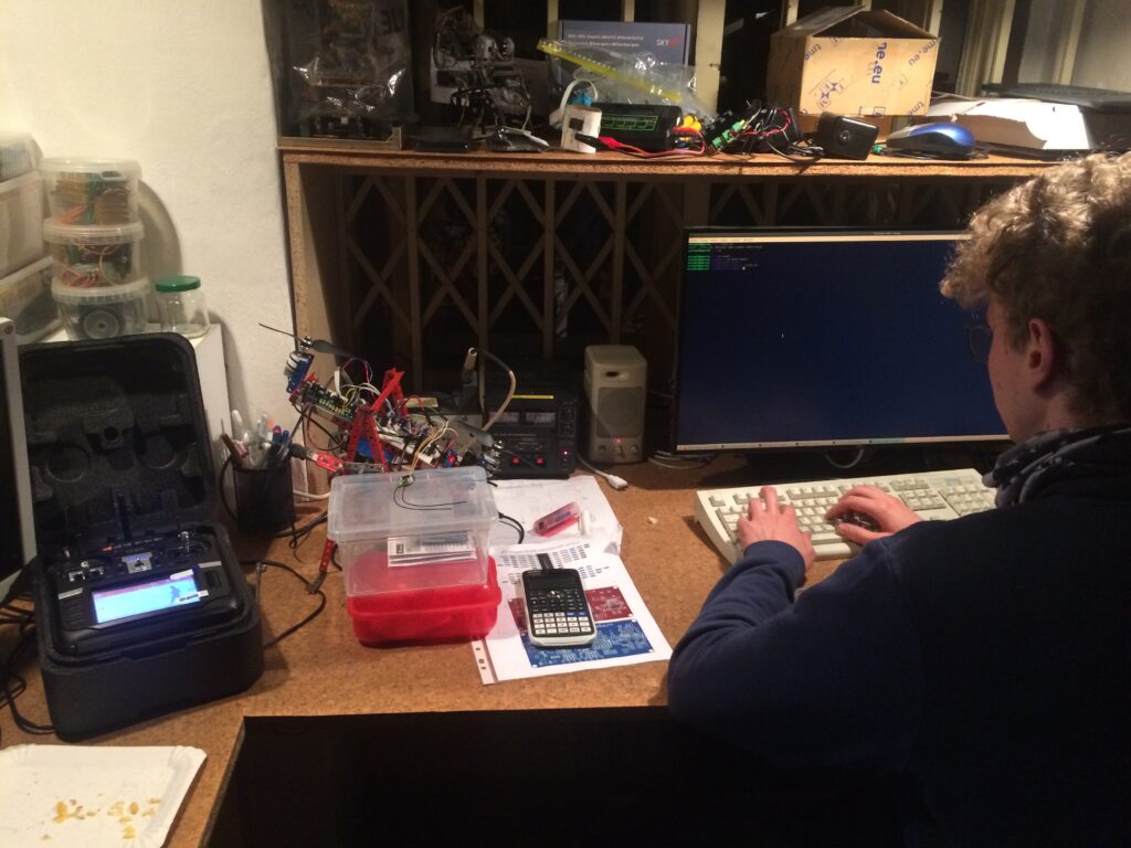 Student píše na klávesnici na přeplněném stole s elektronickým zařízením, včetně počítačového monitoru, modelářské vysílačky, elektroniky a nástrojů.
