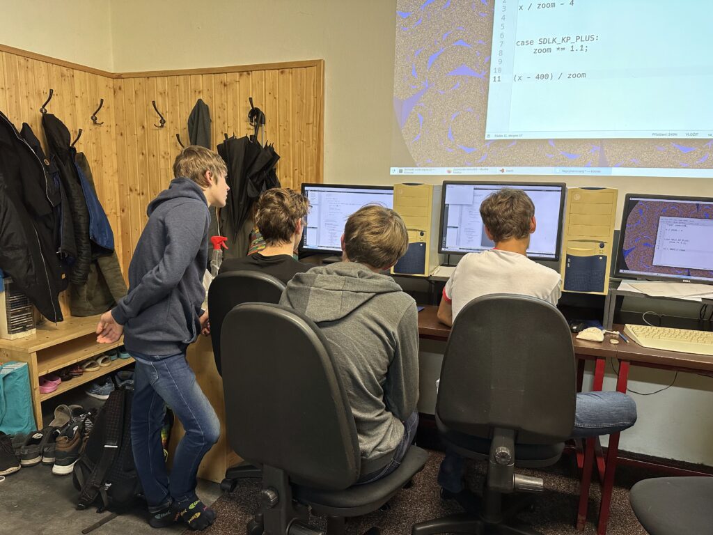 Čtyři studenti sledují počítačový kód na obrazovce, na zdi promítnutý obraz a kód související se zvětšováním Mandelbrotovy množiny a v pozadí kout s věšáky na kabáty.