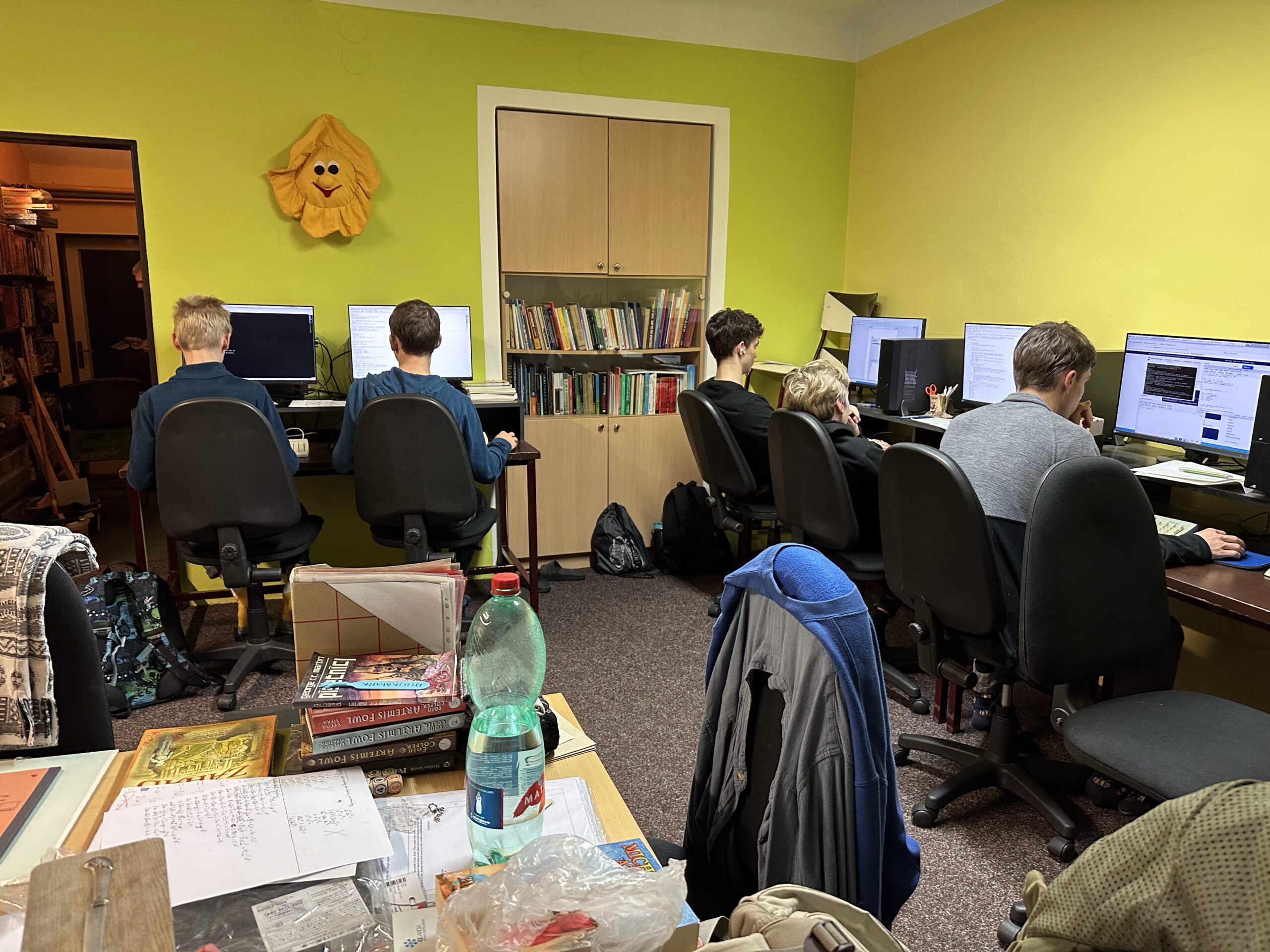Studenti u počítačů v učebně se žlutými stěnami, knihovnou a sluníčkem na zdi.