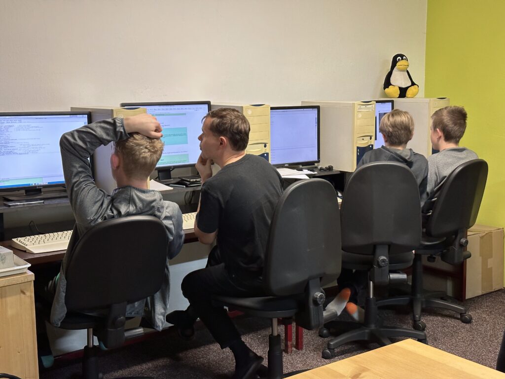 Čtyři studenti u počítačových pracovišť v učebně s plyšovým tučňákem Tuxem na počítačové skříni.