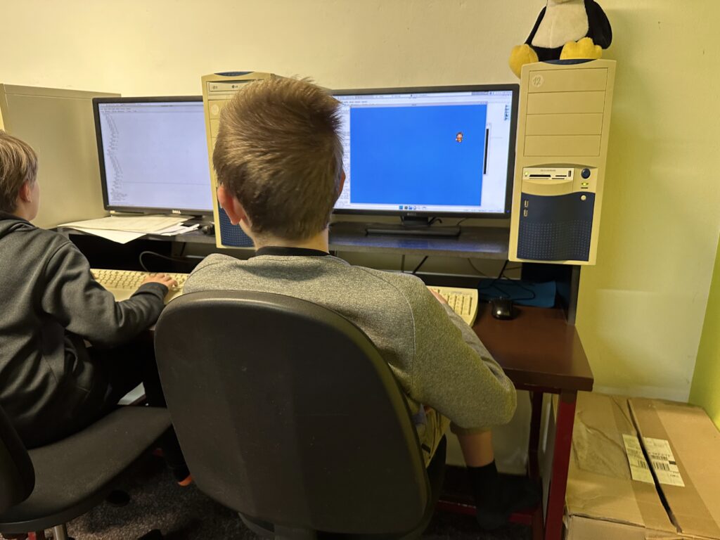Dva studenti u počítačů, na jednom monitoru modrá obrazovka s figurkou, na druhém kód, tučňák Tux na počítačové skříni.