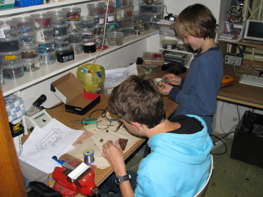 Dva chlapci pracují na elektronických projektech v dílně s nářadím, kontejnery na součástky a schématy obvodů na stole.