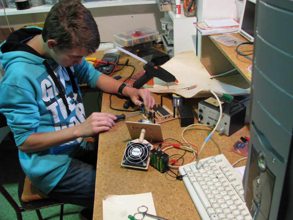 Kluk v modré mikině používá nástroje pro montáž elektroniky na zaplněném pracovním stole s elektronickými deskami, dráty a počítačovým vybavením.