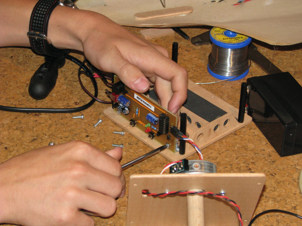 Ruce studenta montují elektronický obvod se šroubovákem, okolo součástky, šrouby a nářadí.