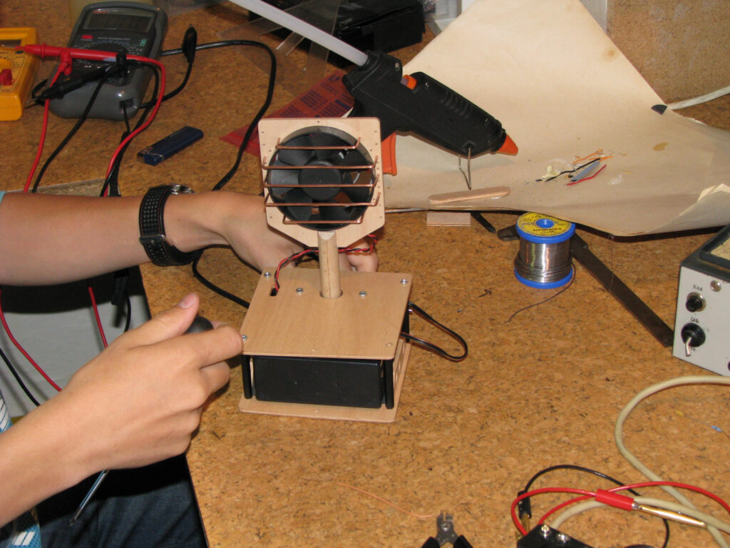 Ruce kluka pracujího na elektronickém zařízení s ventilátorem, dráty a nářadím na zaplněném pracovním stole.