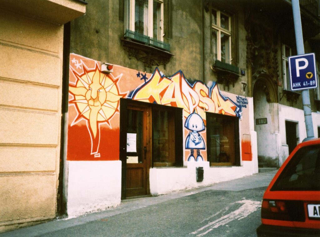 Stará fotografie vstupu do Kapsy z ulice s barevným graffiti na fasádě zobrazující umělecky ztvárněné slunce s lvím obličejem, stylizovaným textem a obrázkem postavy robota.