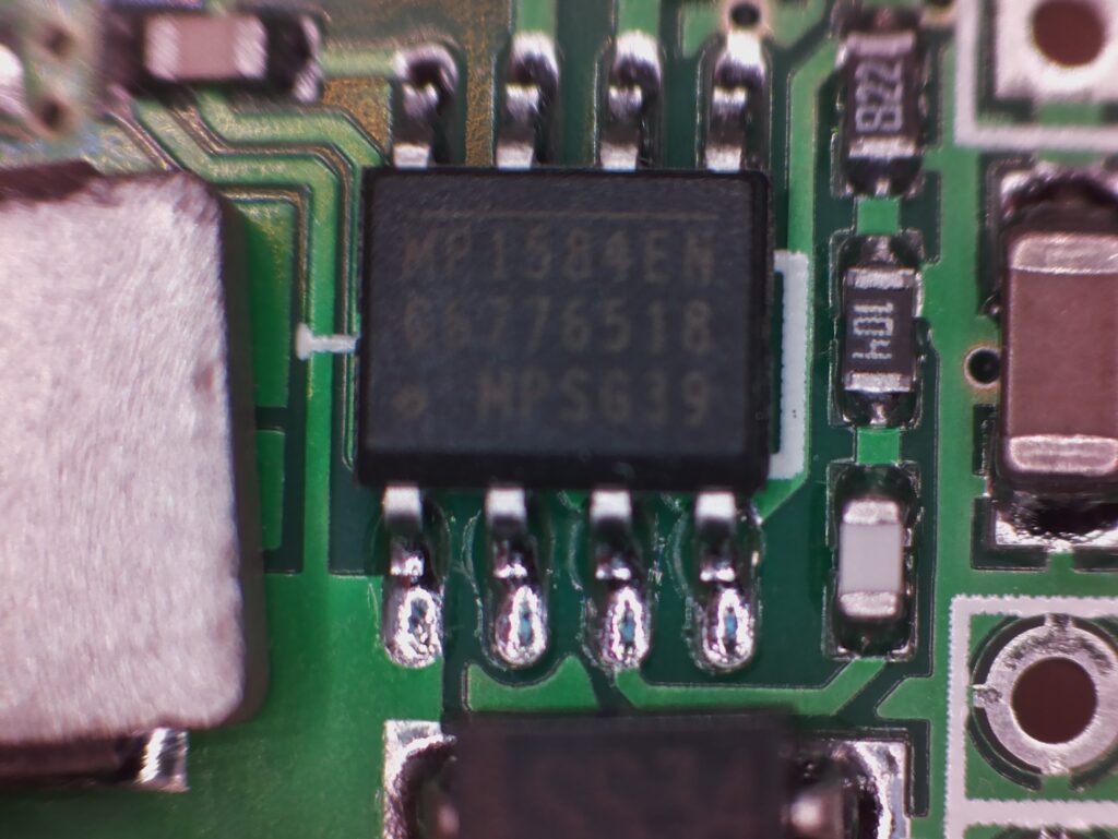 Mikroskopický pohled na integrovaný obvod MP1584EN na zelené desce tištěných obvodů s okolními elektronickými součástmi.